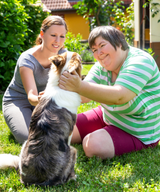 An adult with an adult caregiver pets a dog outside