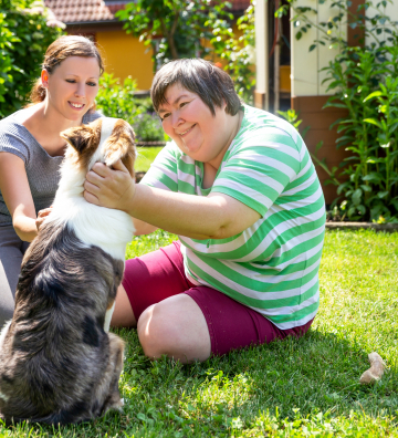 An adult with an adult caregiver pets a dog outside
