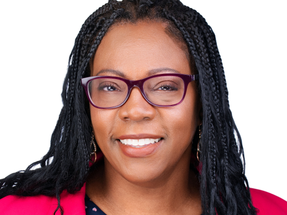 An adult with long box braids with curly ends and medium brown skin smiles in a posed headshot. She is wearing eyeglasses and a pink blazer.