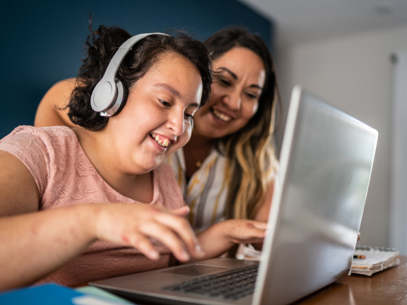 A young adult with headphones types on a laptop while an adult looks over their shoulder smiling.