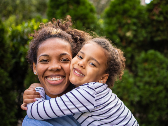 a person with brown hair holds a child and smiles at the camera in front of some trees.