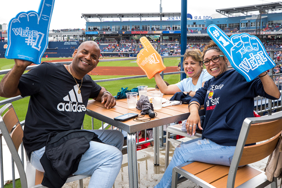 Three smiling Advocates staff members sitting at a table at a WooSox game. They each have a foam finger in Advocates orange and blue that says "We're number one."