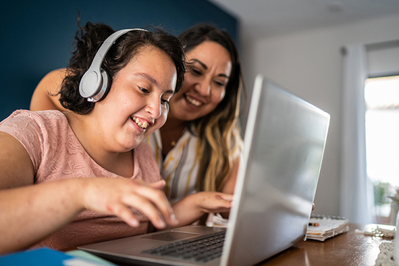 Young woman wearing headphones working on a laptop while her mother stands by