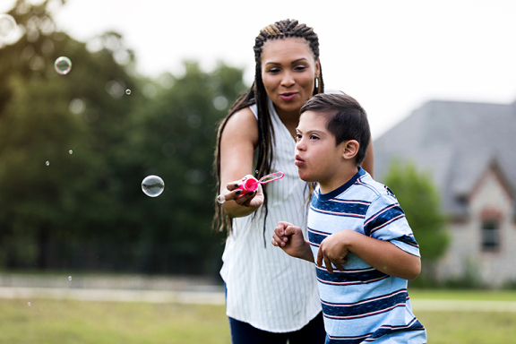 Adult woman holding bubble wand for a child with down syndrome who is blowing bubbles.