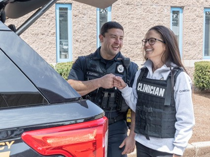 A clinician and an officer smile and fist bump behind an open SUV trunk.