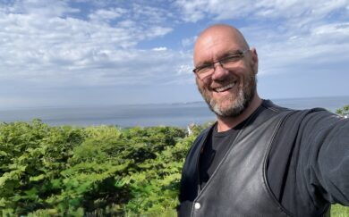 A light-skinned, bald adult with facial hair and glasses takes a smiling selfie atop a mountain or hill.