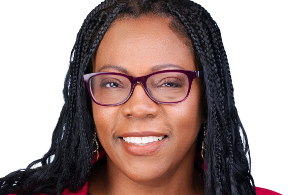 An adult with long box braids with curly ends and medium brown skin smiles in a posed headshot. She is wearing eyeglasses and a pink blazer.