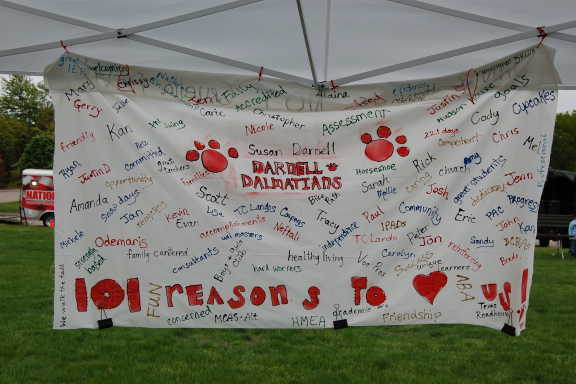 A large white banner hangs outdoors under a tent. It is hand-painted with names and memories from Darnell School.