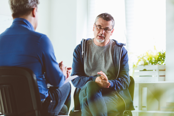 Middle-aged person with glasses and gray goatee sits across from a therapist