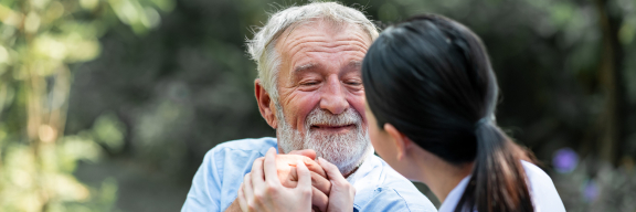 Older person sits in wheelchair, smiling and grasping hands with a younger caregiver. They are outdoors.