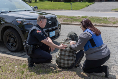 An officer and a clinician comfort a person sitting on a curb in front of a police cruiser