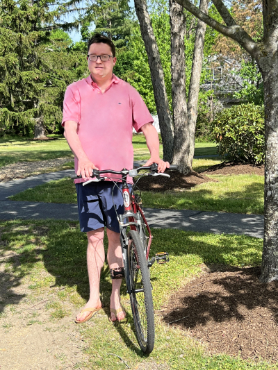 Jack, a young man, standing next to a tree with his bike. He is wearing a pink polo shirt, shorts, and glasses.