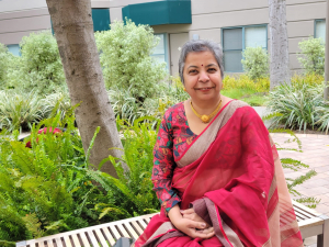 A person with light brown skin and short grey hair is wearing a pinkish red saree. She is smiling at the camera outdoors.