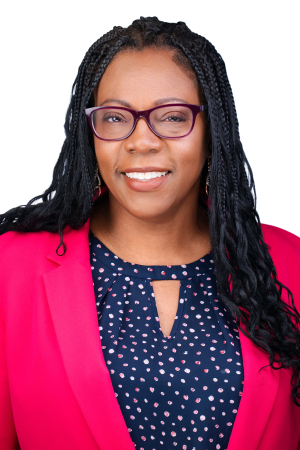 An adult with long box braids with curly ends and medium brown skin smiles in a posed headshot. She is wearing eyeglasses and a pink blazer.