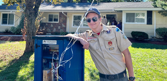 Eagle Scout Jim Kemp posing with one of the Little Free Libraries he and his troop installed at Advocates residential programs. There are colorful balloons tied to the library.