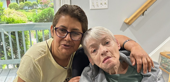 A middle aged adult with short hair and glasses embraces a senior adult using a wheelchair. They are posing for a picture in a home's hallway.