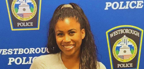 A smiling brown-skinned adult with long dark hair stands in front of a Westborough Police Department step-and-repeat background