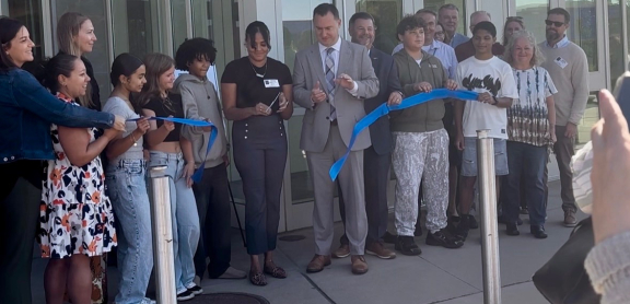 A group of students and adults cut a blue ribbon in front of a school building