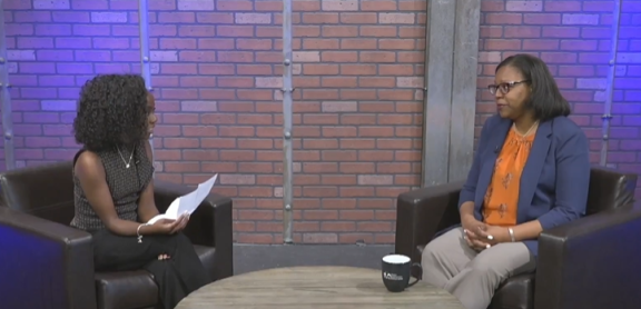 An interview between two Black adults with shoulder length hair on a television set/stage.