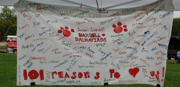 A large white banner hangs outdoors under a tent. It is hand-painted with names and memories from Darnell School.